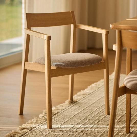 Ljus ekfärgad matbordsstol med armstöd och beige tygsits på en juteliknande matta i ett ljust matrum.