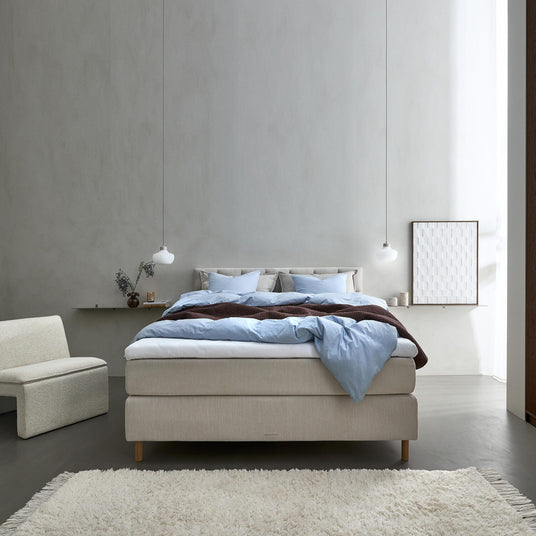 Minimalistiskt sovrum med beige kontinentalsäng, ljusblått bäddset, två hängande vita pendellampor och en mjuk ljus matta i lugn dagsbelysning.