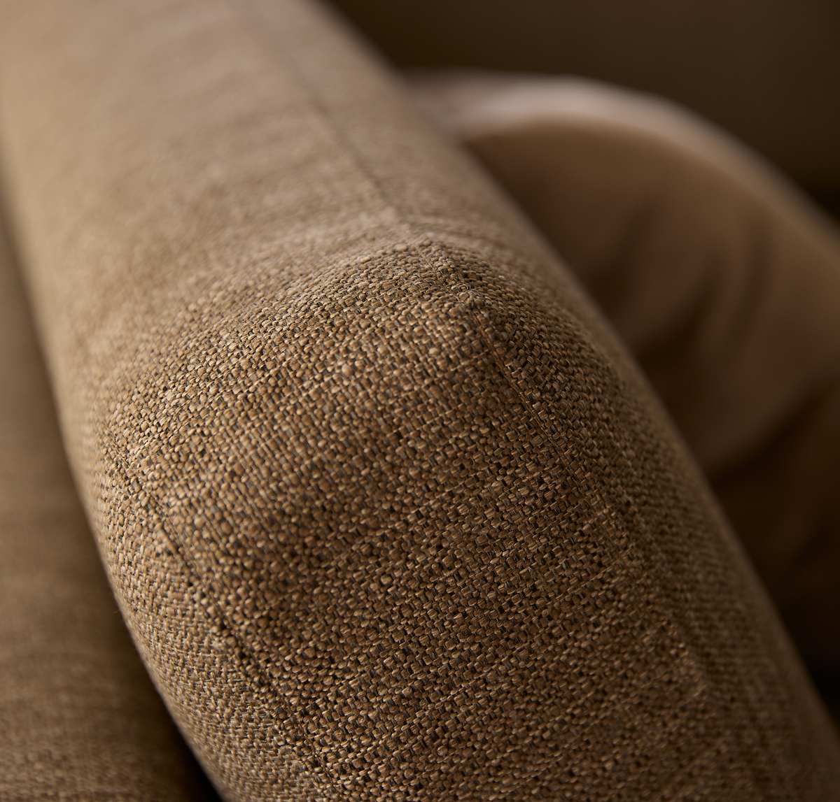 Närbild av soffans armlän i brun vävd textil med mjukt rundad form och en kudde i bakgrunden.