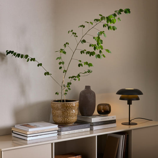 Beigefärgad sideboard med grön krukväxt, keramikvas, tänd ljuskopp och svart bordslampa ovanpå staplade böcker som The Kinfolk Home och The Home Upgrade mot en neutral vägg.