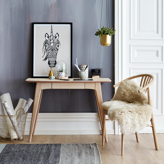 Ljus skandinavisk entré med konsolbord i ljust trä, stol med fårskinn, hängande guldfärgad kruka och en inramad svartvit illustration av en enhörning.