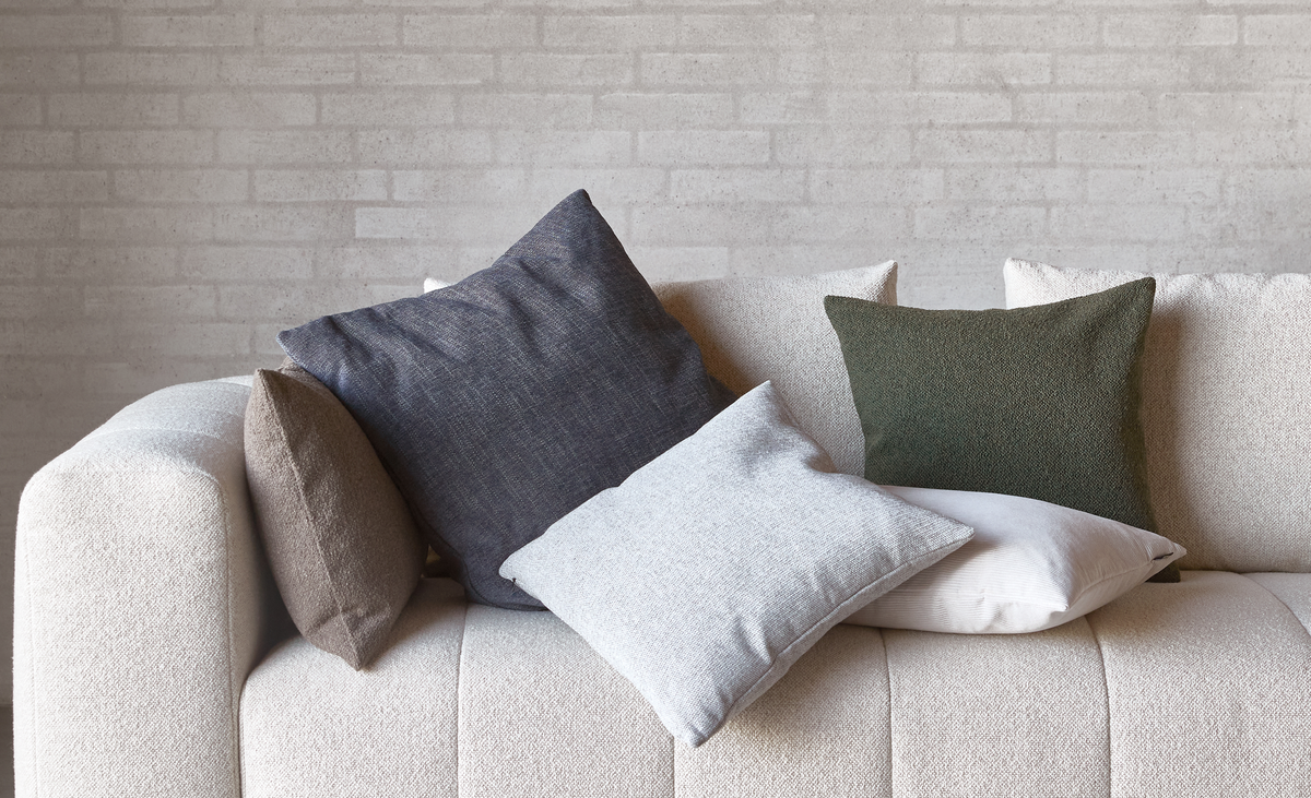Beige soffa med flera prydnadskuddar i grått, grönt, vitt och brunt mot en ljus tegelvägg.