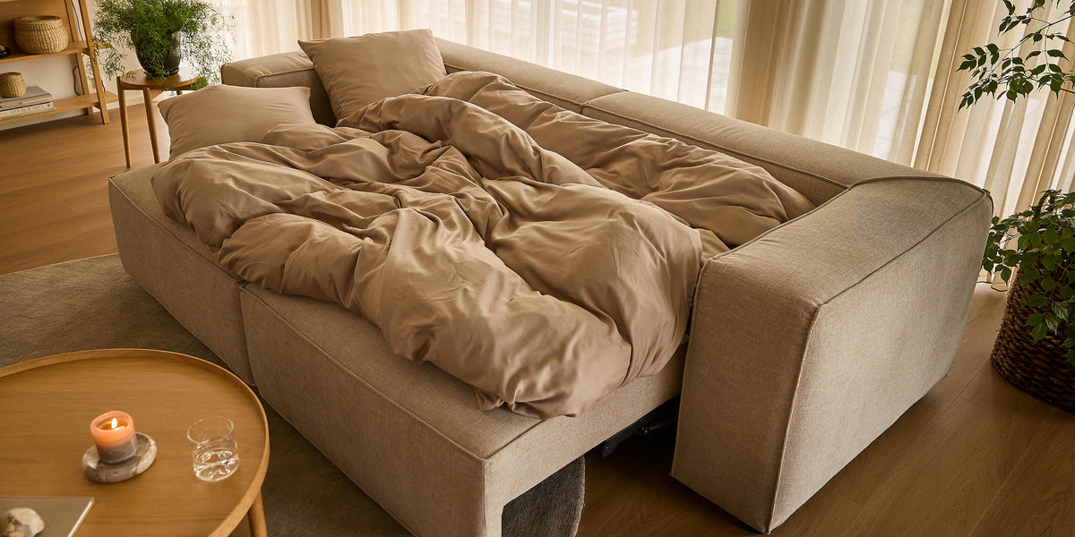 Beige bäddsoffa uppfälld med sandfärgat påslakan i ett ljust vardagsrum med skira gardiner, runt träbord med tändt ljus och gröna växter.