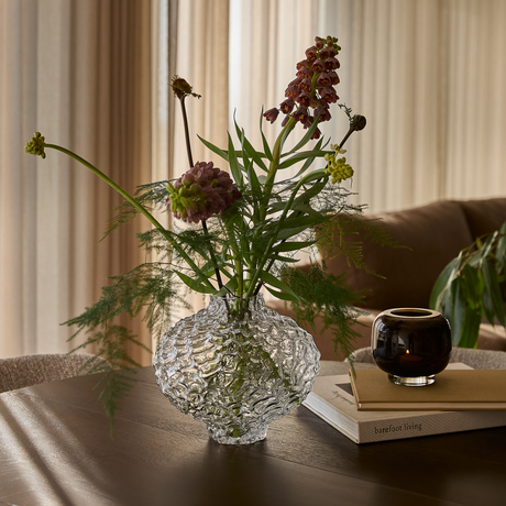 Vågstruktur glasvas med blommor på träbord vid soffa; rund mörk ljuslykta på böcker, varav en med ”barefoot living”, mjuka gardiner i bakgrunden.