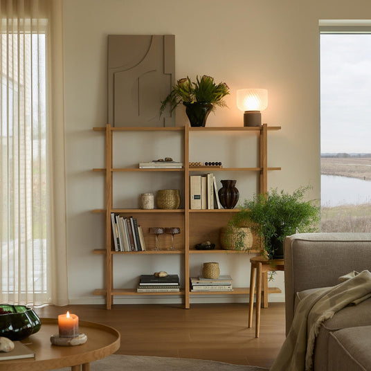 Ljus och varm vardagsrumsbild med en ekbokhylla stylad med böcker, korgar, växter och en glödande bordslampa; tänd värmeljus på runt soffbord och neutral soffa vid stort fönster.