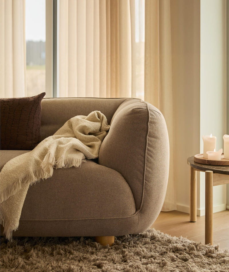 Närbild av en varm, neutral hörna i vardagsrummet: brun soffa med krämfärgad pläd och kudde, lång luggmatta och litet träbord med tända ljus framför beige gardiner i dagsljus.