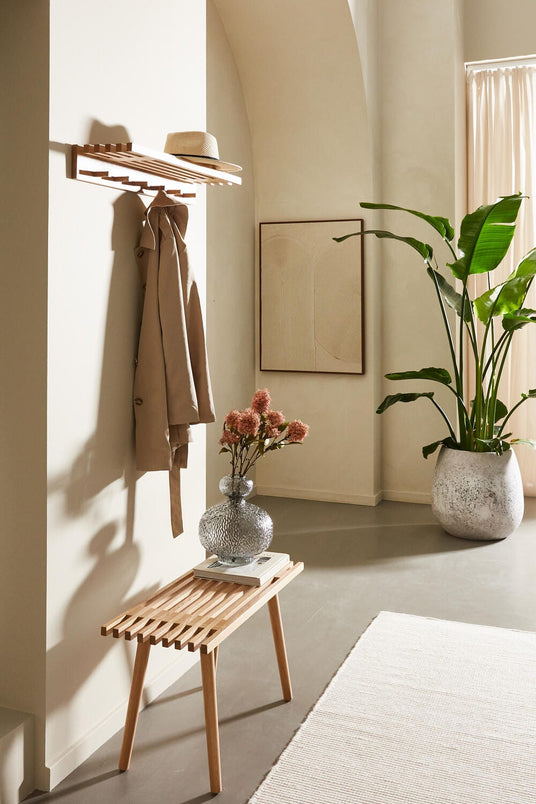 Ljus hall med träbänk, väggmonterad hatthylla med beige kappa och stråhatt, stor grön växt och glasvas med blommor.