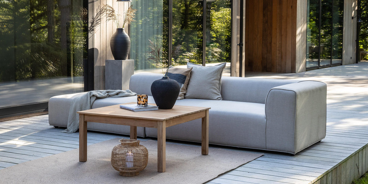 Modern uteplats med ljusgrå soffa, träbord och vaser framför ett trähus med stora glaspartier i dagsljus.