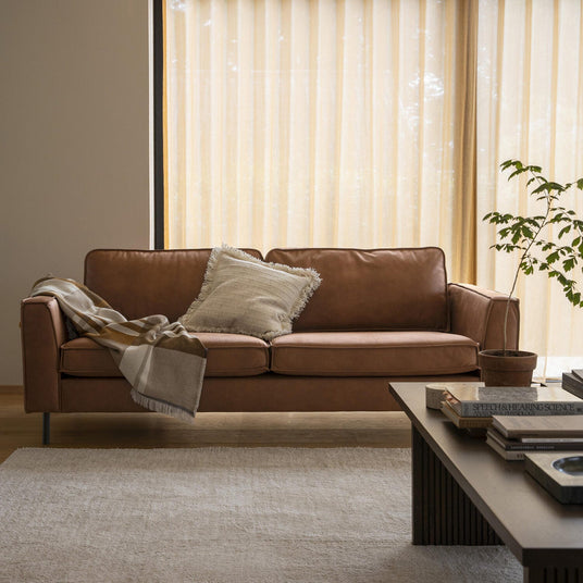 Ljust vardagsrum med brun lädersoffa, franskantad kudde och filt, beige gardiner, en krukväxt och en ljus matta på trägolv i dagsljus.