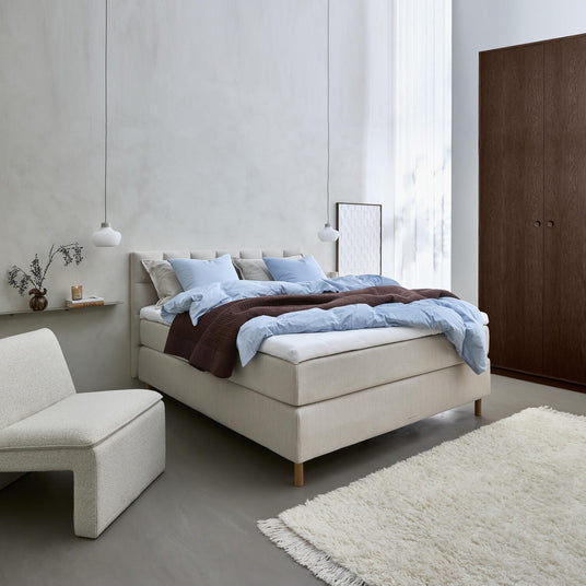 Ljust skandinaviskt sovrum med beige stoppad säng, ljusblått bäddset och brun pläd, två pendellampor, låg fåtölj, vas med kvistar på hylla, skira gardiner, vit ryamatta och mörk trägarderob.