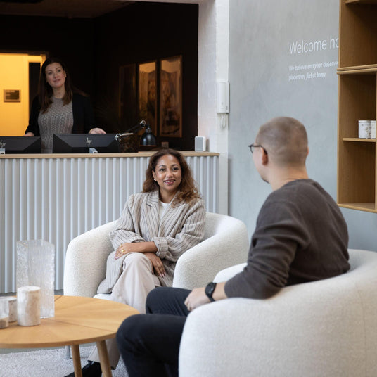 Modern showroom-reception med två personer i vita fåtöljer och runt träbord med glasvaser; på väggen står “Welcome home – We believe everyone deserves a place they feel at home”.