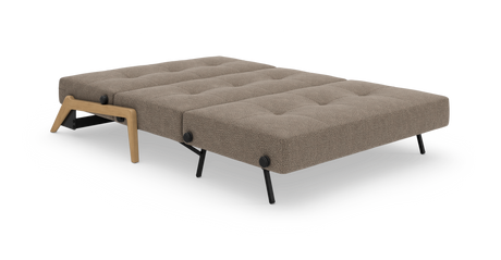 Bäddsoffa i utbäddat läge, beige/taupe tyg med knapptuftad yta, svarta metallben och träben i ek-look, frilagd mot vit bakgrund.