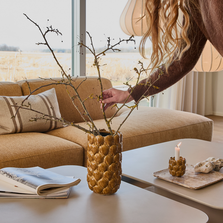 Hand som arrangerar grenar i en brun, strukturerad vas på soffbord; beige soffa, tändt stearinljus og öppnad bok i ett ljust vardagsrum.