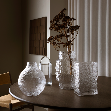 Samling av strukturerade glasvaser på ett mörkt runt bord med torkad kvist i en vas, i en neutral inredning med gardiner.