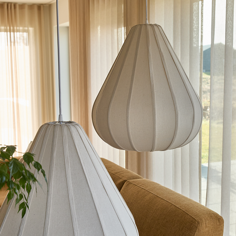 Närbild på två beige tygklädda pendellampor ovanför en senapsgul soffa framför ljusa gardiner i ett vardagsrum.