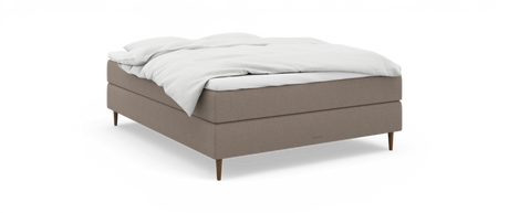 Beige stoppad kontinentalsäng med vit bäddning och avsmalnande träben, sedd snett framifrån; liten logotyp ”necturne” på sängkanten.