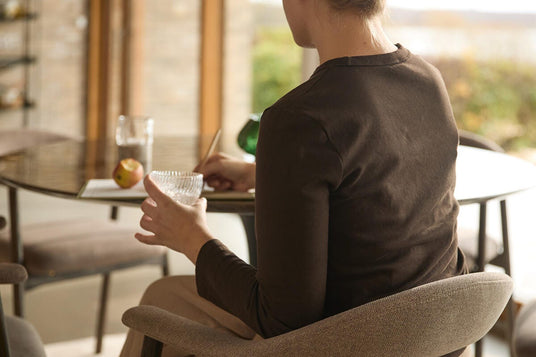 Person sitter på en stoppad spisestol vid ett runt matbord, håller ett glas och antecknar i ett ljust, luftigt matrum.
