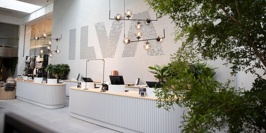Modern butikskassa med hängande lampor, gröna växter och stor ILVA‑logga på väggen.