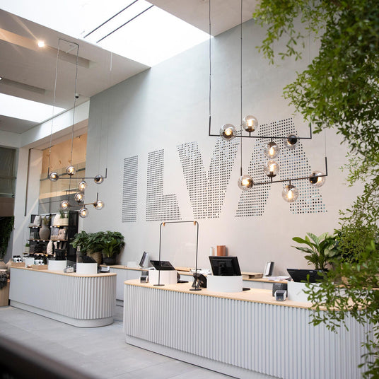 Modern butikskassa med hängande lampor, gröna växter och stor ILVA‑logga på väggen.