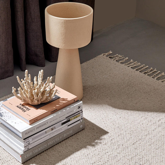 En stapel designböcker, bl.a. “THE DESIGN HOTELS BOOK”, “PERFECT IMPERFECT” och “Creative”, toppad med en korallskulptur bredvid en beige texturerad bordslampa på en neutral vävd matta med frans; mjukt dagsljus och mörka gardiner i bakgrunden.