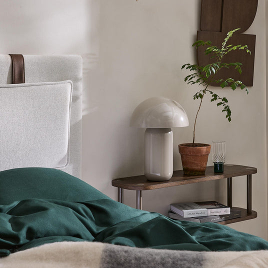 Sovrumsnattduksbord med svampformad bordslampa, terrakottakruka med liten planta och böcker; säng med gröna textilier i förgrunden.