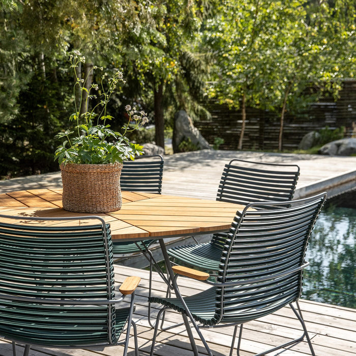 Runt teakbord med gröna metallstolar på en träaltan vid en pool, med en korgkruka med grön växt i mitten i ett soligt trädgårdsläge.