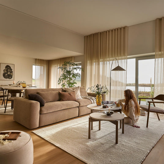 Ljus och varm vardagsrumsmiljö med beige soffa, helhöga skira gardiner och två runda soffbord; en person sitter på mattan med kopp och bok.