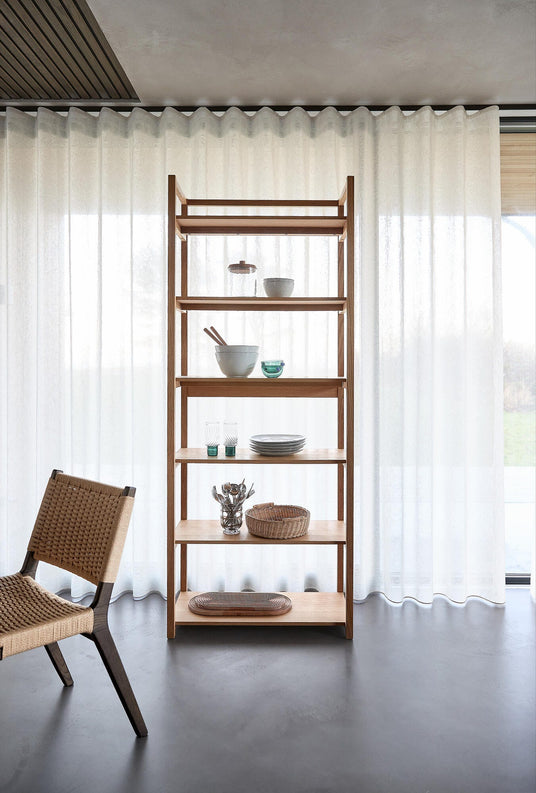 Öppen hylla i ljust trä med keramikskålar, glas och korg framför ljusa skira gardiner; flätad stol i vänsterkant.