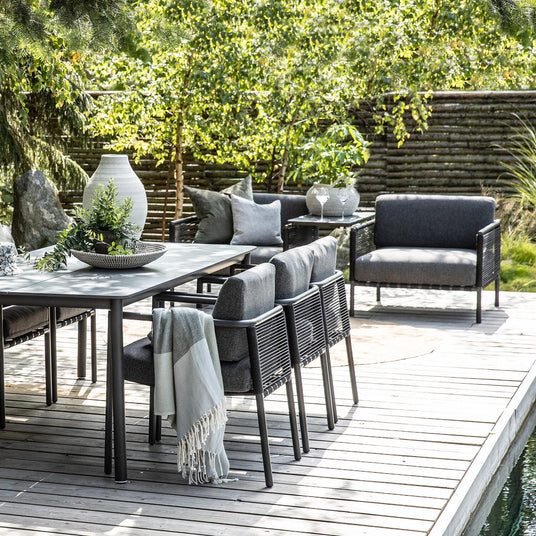 Utemöbler på träterrass: svart matbord med grå stoppade stolar, loungefåtölj och soffa; bordet dekorerat med vit vas, skål med gröna kvistar och en ljus pläd, omgiven av frodig trädgård.