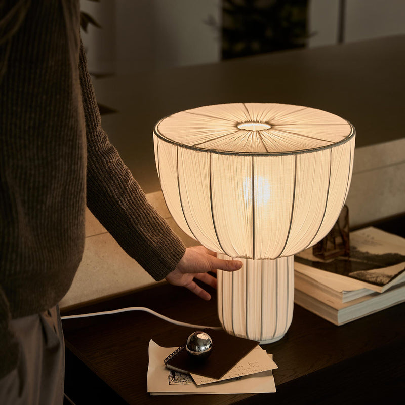 Hand som justerar en varmglödande bordslampa med plisserad skärm på ett mörkt sidobord med böcker.
