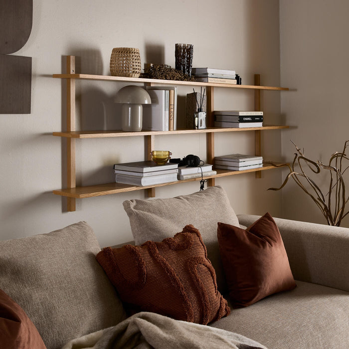 Väggmonterad hylla i ljus ek över beige soffa med kuddar; böcker som Chanel och New Nordic Houses, liten bordslampa och flätad korg på hyllplanen.