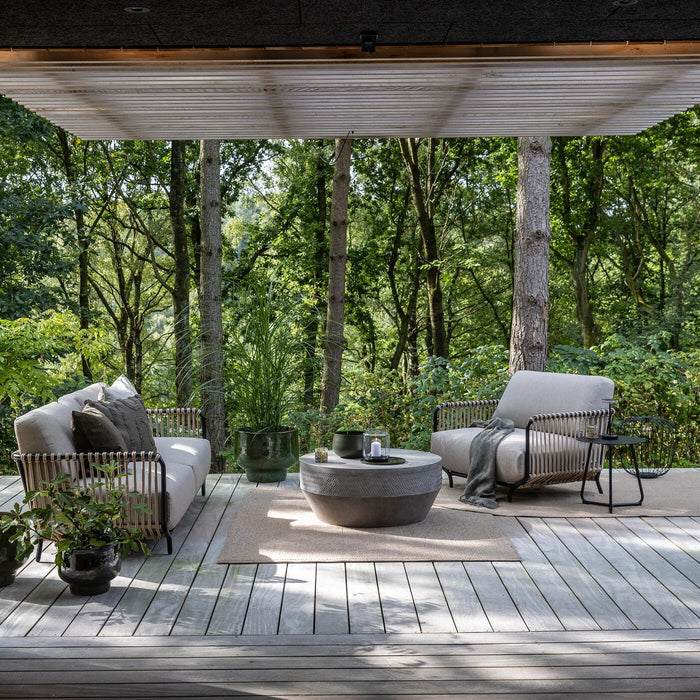 Uteplats på trädäck i skog med loungesoffa och fåtölj, runt betongbord, mattor, krukväxter och staplad ved under pergola.