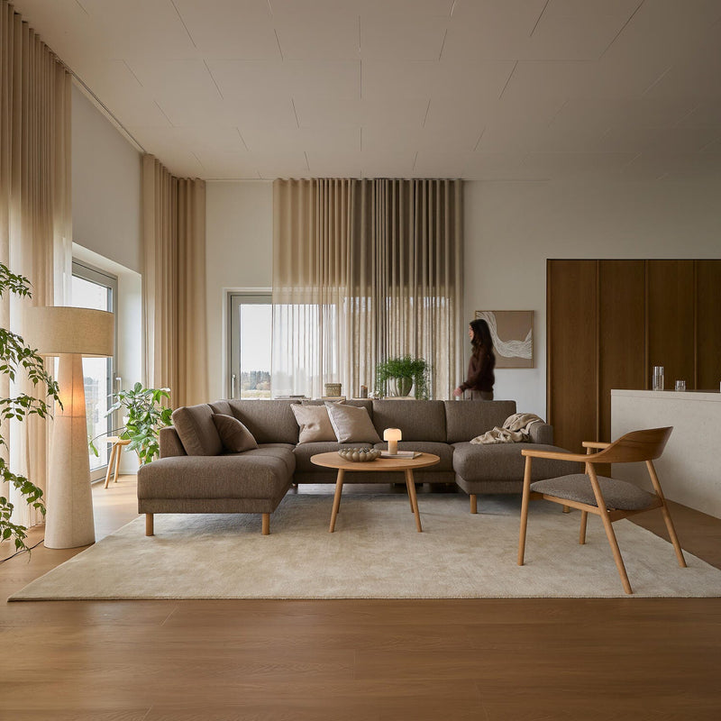 Ljust skandinaviskt vardagsrum med beige hörnsoffa, träfåtölj och soffbord på ljus matta, skira gardiner, gröna växter och ett tänt ljus.
