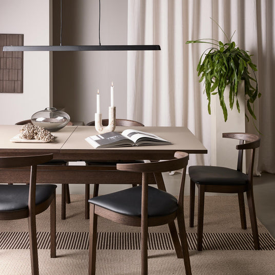 Minimalistisk matplats med mörka trästolar, beige bord, pendellampa, tända ljus, glasvas, öppen bok och hög grön växt framför ljusa gardiner.