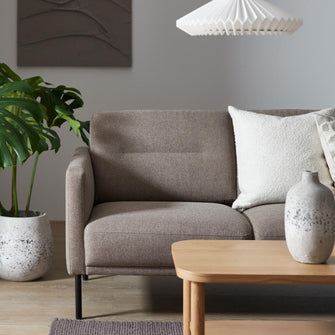Beige soffa med vita kuddar, ljus träsoffbord, keramikvas, plisserad vit pendellampa och grön monstera i ett vardagsrum.