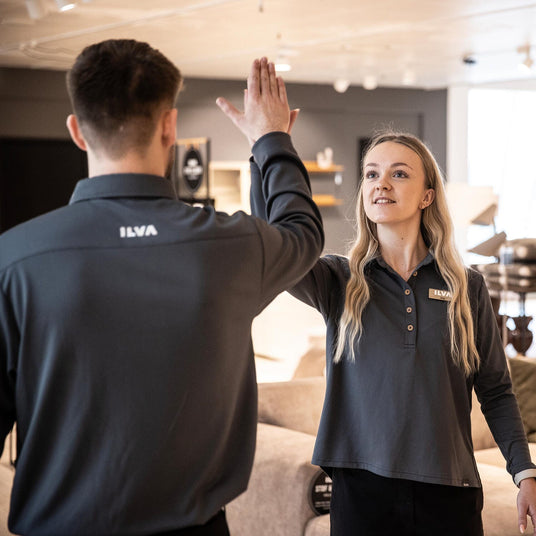 Två ILVA‑medarbetare i mörka uniformer gör en high‑five i en möbelbutik/showroom.
