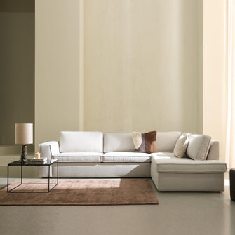 Ljus, minimalistisk vardagsrumsscen med beige modulsoffa med schäslong, glasbord med bordslampa och ljus, brun matta och mjukt dagsljus genom ljusa gardiner.