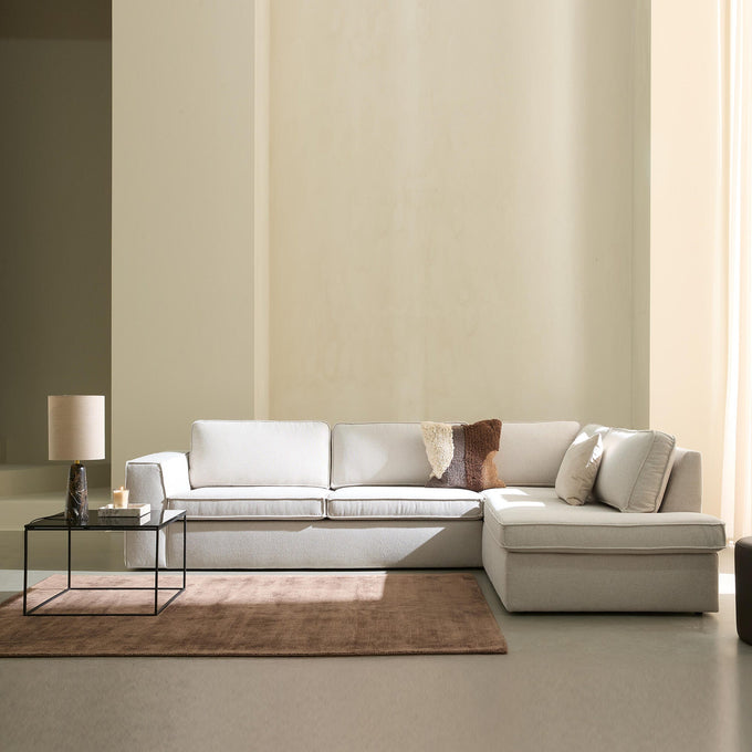 Ljus, minimalistisk vardagsrumsscen med beige modulsoffa med schäslong, glasbord med bordslampa och ljus, brun matta och mjukt dagsljus genom ljusa gardiner.
