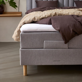 Grå stoppad säng med vertikal ribbad gavl, brunt bäddset och beige quilt, träben och märkning nocturne; bredvid en vägghylla med vas, grön växt och vägglampa.