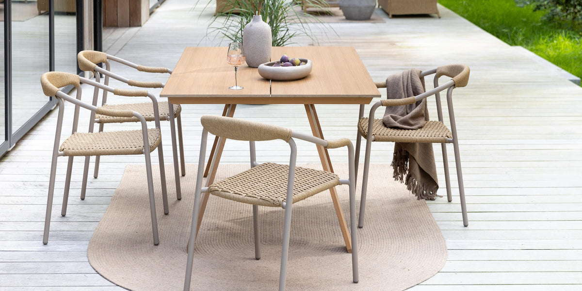 Uteservering på trädäck: fyrkantigt träbord med flätade stolar, beige utomhusmatta, vas med blommor, skål med frukt och vinglas; lounge-möbler och grön trädgård i bakgrunden.