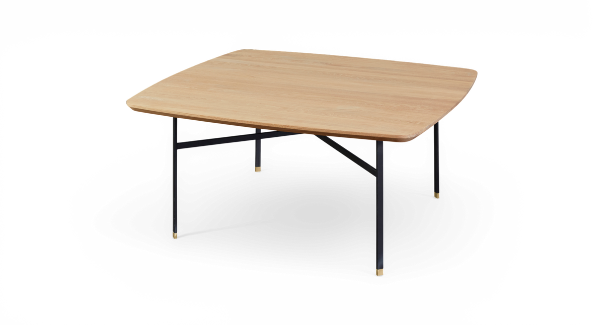 Lågt soffbord med asymmetrisk kvadratisk bordsskiva i ljus ek och svarta metallben med små mässingsdetaljer, frilagd mot vit bakgrund.