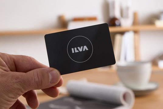 Hand som håller ett svart kort med ILVA‑logga, med suddig heminredning, kopp och tidning på bord i bakgrunden.