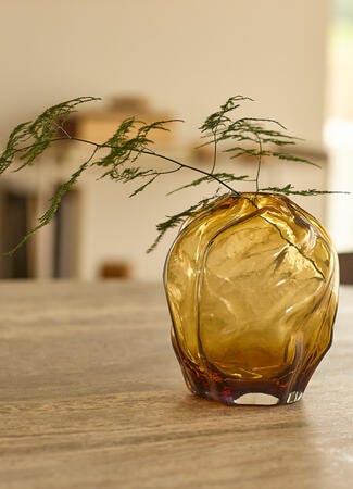 Bärnstensfärgad glasvas med tunna gröna kvistar på ett träbord i ett ljust vardagsrum.