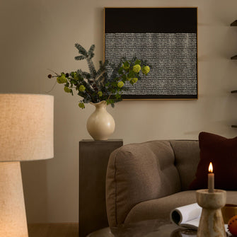 Varmt inredd hörna i vardagsrum med beige soffa, vit keramikvas med gröna kvistar på piedestal, bordslampa och tänd ljus, abstrakt svartvit tavla på väggen.