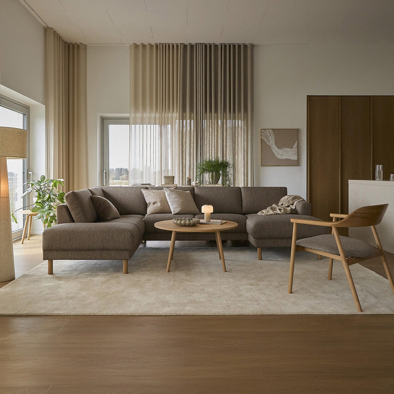 Ljus och varm vardagsrumsinteriör med beige gardiner, brun modulsoffa, runt träbord med tändt ljus och stor ljus matta.