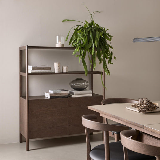 Lugn matplats i skandinavisk stil med mörk träsideboard och öppna hyllor med böcker, ljus, glasvas och hängande grön växt intill bord och stolar; en bok har titeln Dior.