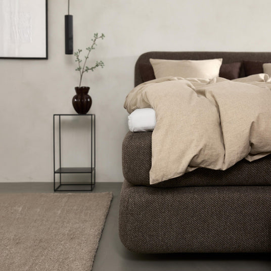 Minimalistiskt sovrum med brun stoppad säng, beige linnesängkläder, svart metall-sängbord med vas och kvist, hängande pendellampa och neutral matta.