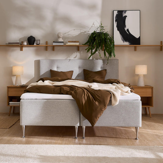 Sovrum med ljusgrå kontinentalsäng, brunt bäddset och beige stickad pläd; två trä‑sängbord med bordslampor; lång trähylla med böcker (BOSS), vas och dekor, väggkonst och hängande grön växt i mjukt dagsljus.