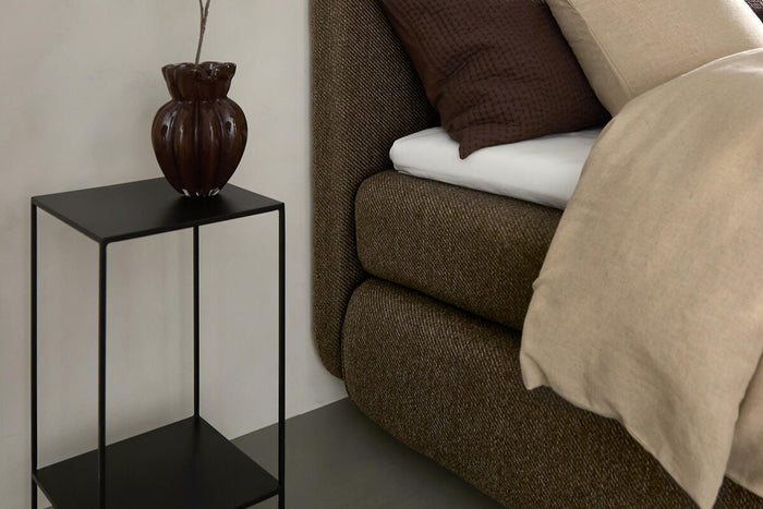 Sovrumsnärbild med brun stoppad säng, beige och bruna kuddar, svart metall-sidobord med brun vas och kvist samt en hängande svart pendellampa.