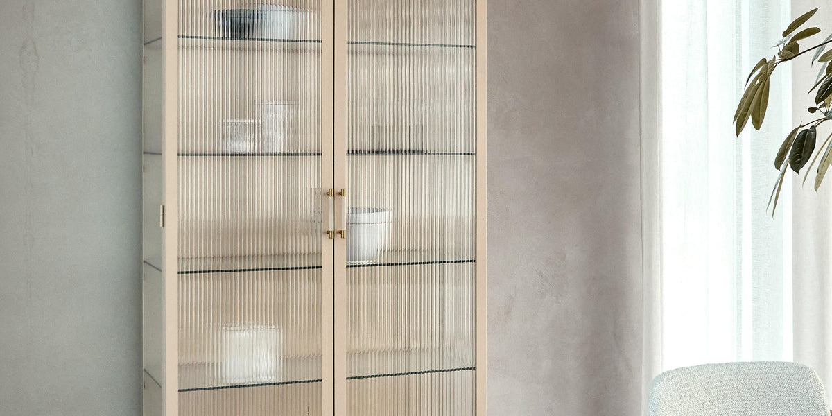 Beige vitrinskåp med räfflat glas och smala ben i ett ljust matrum; rund träbordsskiva och tunn gardin i dagsljus.